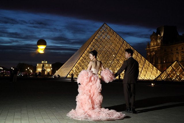 A couple pose for photographs in front of the Louvre Pyramid, designed by Chinese-US architect Ieoh Ming Pei, as the Paris 2024 Olympic cauldron, designed by French designer Mathieu Lehanneur, flies in the background, in central Paris on September 8, 2025. (Photo by Ludovic Marin/AFP Photo)