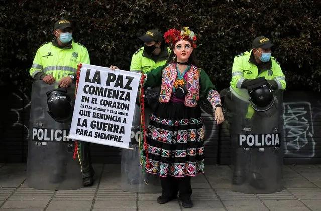 A woman dressed in a traditional Mexican dress and mask holds a sign that reads in Spanish “Peace begins in the heart of every man and woman to renounce war forever”, as police stand guard on the sidelines of an anti-government march in Bogota, Colombia, Wednesday, May 19, 2021. Colombians have taken to the streets for weeks across the country after the government proposed tax increases on public services, fuel, wages and pensions. (Photo by Ivan Valencia/AP Photo)