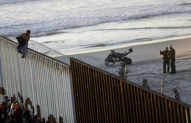 A Central American migrant sits on top of the border structure separating Mexico, left, and the United States, right, as U.S. Border Patrol agents look on, right, Wednesday, November 14, 2018, seen from Tijuana, Mexico. Migrants in a caravan of Central Americans scrambled to reach the U.S. border, catching rides on buses and trucks for hundreds of miles in the last leg of their journey Wednesday as the first sizable groups began arriving in the border city of Tijuana. (Photo by Gregory Bull/AP Photo)