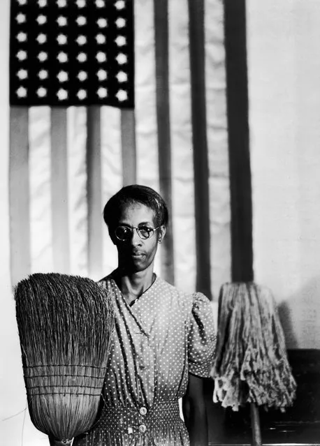 Ella Watson, a government charwoman, in Washington DC, August 1942. She is wearing a polka dot smock, standing in front of an American flag, and holding a broom in one hand and a mop in the other. (Photo by Gordon Parks/FSA/Getty Images)