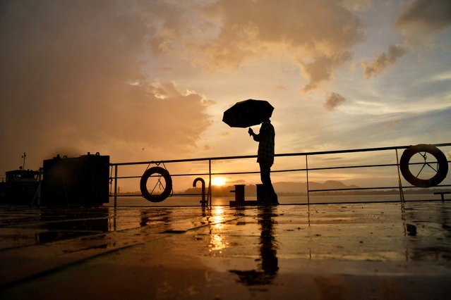 A man holding an umbrella is silhouetted against the setting sun as it rains along the banks of river Brahmaputra in Guwahati on July 25, 2024. (Photo by Biju Boro/AFP Photo)