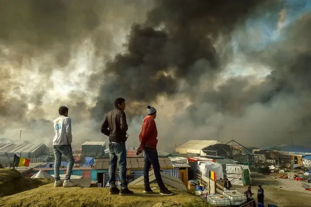Migrants stand on a hill overlooking the “Jungle” migrant camp in Calais, northern France, as smoke rises on October 26, 2016 during a massive operation to clear the squalid settlement where 6,000-8,000 people have been living in dire conditions. Fresh fires broke out on October 26 in the “Jungle” migrant camp on the second day of operations to dismantle the squalid settlement in northern France. Smoke billowed over the sprawling camp near Calais for a second day amid fears that abandoned gas cylinders could explode. (Photo by Philippe Huguen/AFP Photo)