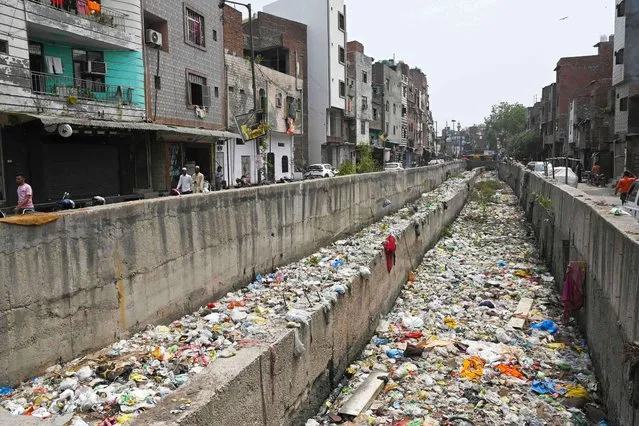 In this photograph taken on April 26, 2023, people walk along a street next to a sewage canal filled with garbage at Seelampur neighbourhood in New Delhi. Out of the 72 billion litres of sewage currently generated in Indian urban centres every day, 45 billion litres – equal to 18,000 Olympic-sized swimming pools – isn't treated, according to government figures for 2020-21. (Photo by Arun Sankar/AFP Photo)