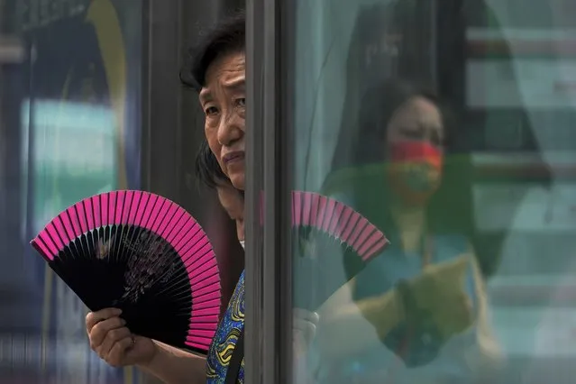 A woman fans herself at an advertisement boards on a bus stand reflecting a masked commuter as they wait for buses in Beijing, Tuesday, July 5, 2022. (Photo by Andy Wong/AP Photo)