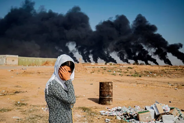 A woman covers her face as she stands along the side of a road on the outskirts of the town of Tal Tamr near the Syrian Kurdish town of Ras al-Ain along the border with Turkey in the northeastern Hassakeh province on October 16, 2019, with the smoke plumes of tire fires billowing in the background to decrease visibility for Turkish warplanes that are part of operation “Peace Spring”. (Photo by Delil Souleiman/AFP Photo)
