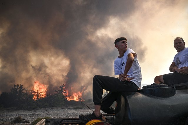 Men sit on the back of their pick-up truck as wildfires rage in the background close to the village of Vati on the Greek island of Rhodes on July 25, 2023. Some 30,000 people fled the flames on Rhodes at the weekend, the country's largest-ever wildfire evacuation as the prime minister warned that the heat-battered nation was “at war” with several wildfires and spoke of three difficult days ahead. (Photo by Spyros Bakalis/AFP Photo)