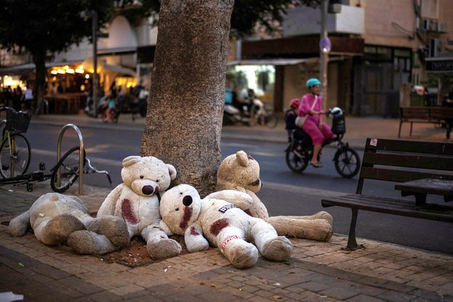 A woman with a child rides a bicycle passing by teddy bears placed by a tree, symbolising hostages kidnapped during the deadly October 7 attack by  Hamas, in Tel Aviv, Israel on May 28, 2024. (Photo by Marko Djurica/Reuters)