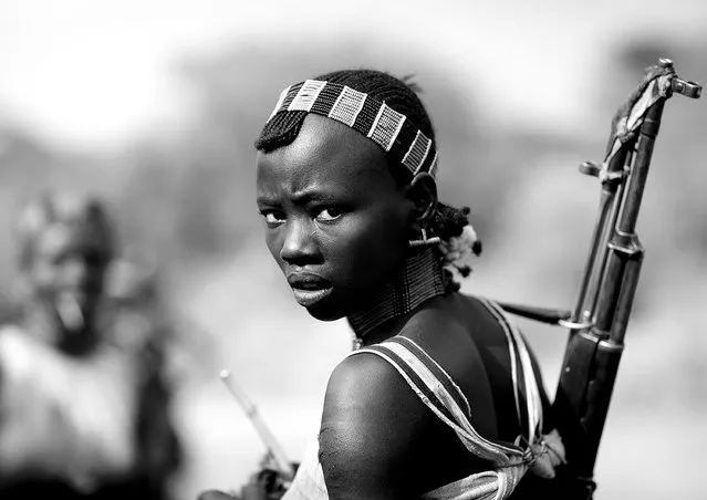 “Hamer girl with gun – Omo Ethiopia. During bull jumping ceremony, Hamer girls act like men, and even more, to show that they do not fear anything. So they drink a lot, and they dance with guns. Very impressive ceremony!
The Hamar is a catlle herder tribe which lives on the Eastern side of the Omo Valley in Southern Ethiopia. Honey collection is their major activity and their cattle is the meaning of their life. There are at least 27 words for the subtle variations of colours and textures of a cattle ! And each man has three names: a human, a goat and a cow name.
The Hamar have very unique rituals such as a bull-leaping ceremony, that a young men has to succeed in order to get married. The cow jumping is an initiation rite of passage for boys coming of age in Hamar tribe. Cows are lined up in a row. The initiate, naked, has to leap on the back of the first cow, then from one bull to another, until he finally reaches the end of the row. He must not fall of the row and must repeat successfully the test four times to have the right to become a husband. While the boys walk on cows, Hamar women accompany him: they jump and sing. Totally committed to their initiated sons, the mothers are whipped to blood, in order to prove their courage and accompany their sons during the test”. (Photo and comment by Eric Lafforgue)