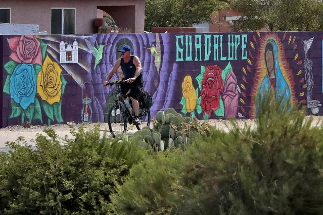 In this Saturday, June 13, 2020, photo, a man cycles past a mural in Guadalupe, Ariz. As the coronavirus spreads deeper across America, it's ravaging through the homes and communities of Latinos from the suburbs of the nation's capital to the farm fields of Florida to the sprawling suburbs of Phoenix and countless communities in between. (Photo by Matt York/AP Photo)
