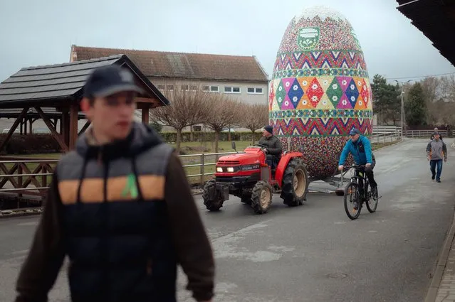 People are seen in front of the world's largest easter egg in Lemesany, Slovakia on April 11, 2022. Local architect and craftsman Jozef Kuruc is the creator of the five-meter tall Easter egg, that has been put on display in the village of Lemesany, Slovakia for the 7th time in a row. The middle-aged architect came up with the idea when he created a tiny, some 50-centimeter tall egg in 2015. The building process of the new, 5.08 meter tall, 3.08 meter wide and nearly 800 kg giant Easter egg lasted for over a year. The body and the shell of the piece are carved out of layers of polyester foam, the outer layer is made out of chicken wire and plaster. The giant egg is fixed onto a metal frame, that also played a key role in its transportation from the workshop to the village's main square. (Photo by Robert Nemeti/Anadolu Agency via Getty Images)
