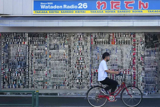 A person cycles past a wall of outdated mobile phones and mock-ups displayed on the exterior of an electronics shop in Tokyo, Japan, Friday, August 18, 2023. The shop owner said his collection started some 20 years ago and now has more than 7,000 mobile phones, drawing attention from passers-by. (Photo by Shuji Kajiyama/AP Photo)