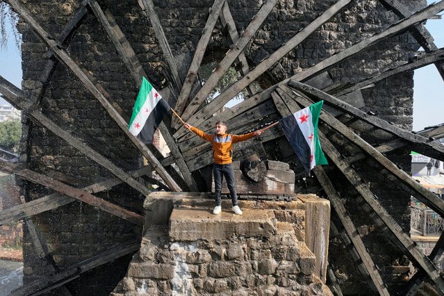 A boy waves the opposition Syrian flag as he stands in one of the water wheels, or norias, in Hama, after anti government fighters captured of the central city, on December 6, 2024. Rebel forces pressing a lightning offensive in Syria aim to overthrow President Bashar al-Assad's rule, their Islamist leader said in an interview published on December 6. In little over a week, the offensive has seen Syria's second city Aleppo and strategically located Hama fall from Assad's control for the first time since the civil war began in 2011. (Photo by Omar Haj Kadour/AFP Photo)