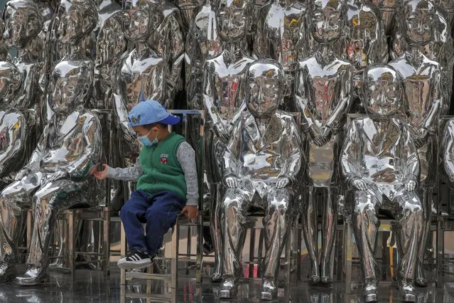 A child wearing a face mask to help curb the spread of the coronavirus tries to hold a statue's hand as he sits in an art installation on display at a shopping mall in Beijing, Sunday, October 11, 2020. Even though the spread of COVID-19 has been all but eradicated in China, the pandemic is still surging across the globe with ever rising death toll. (Photo by Andy Wong/AP Photo)