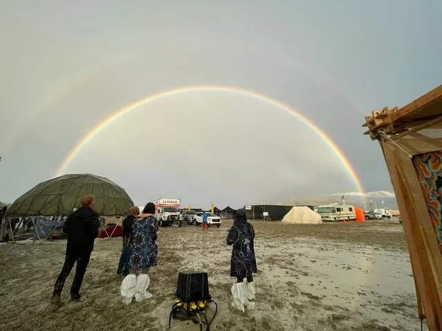 Attendees look at a double rainbow over flooding on a desert plain on September 1, 2023, after heavy rains turned the annual Burning Man festival site in Nevada's Black Rock desert into a mud pit. Tens of thousands of festivalgoers were stranded September 3, in deep mud in the Nevada desert after rain turned the annual Burning Man gathering into a quagmire, with police investigating one death. Video footage showed costume-wearing “burners” struggling across the wet gray-brown site, some using trash bags as makeshift boots, while many vehicles were stuck in the sludge. (Photo by Julie Jammot/AFP Photo)