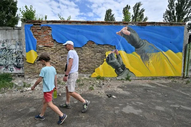 A man and his son walk past a mural by street artist Sasha Korban depicting hands of a military man sewing together parts of the Ukrainian flag, in Kyiv on June 14, 2022. (Photo by Sergei Supinsky/AFP Photo)