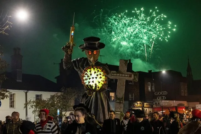 People walk in front of an effigy holding a hypodermic needle, a Covid-19 virus cell and a tombstone as they take part in the parade at the annual Lewes Bonfire Night Celebrations on November 5, 2021 in Lewes, England. The event commemorates the memory of the seventeen Protestant martyrs as Bonfire Societies parade through the narrow streets. The evening comes to an end with the burning of effigies, or “guys” usually representing Guy Fawkes, who died in 1605 after an unsuccessful attempt to blow up The Houses of Parliament, and other topical figures. The annual parade was cancelled last year due to the Covid-19 pandemic. (Photo by Dan Kitwood/Getty Images)