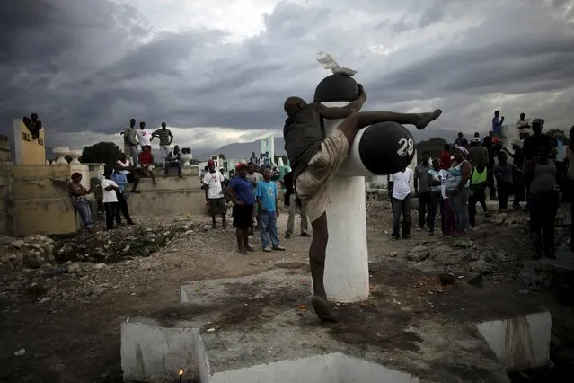 Residents look at a drunk man as he climbs on a Baron Samdi cross, as they wait for the start of a voodoo ceremony, at the cemetery of Croix des Bouquets, on the outskirts of Port-au-Prince, Haiti, November 2, 2015. (Photo by Andres Martinez Casares/Reuters)