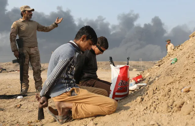 An Iraqi soldier stands next to detained men accused of being Islamic State fighters, at a check point in Qayyara, south of Mosul, Iraq October 27, 2016. Smoke in the background is from burning oilfields set ablazed by Islamic State fighters. (Photo by Goran Tomasevic/Reuters)