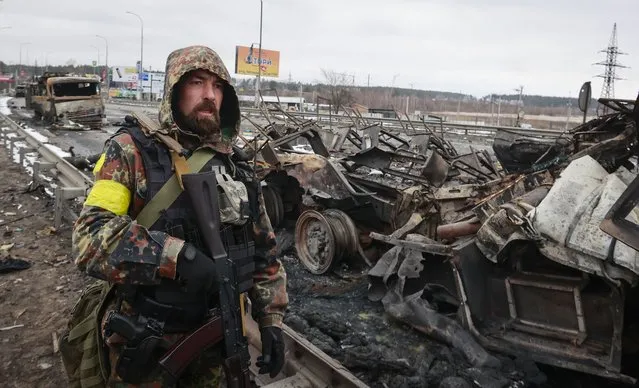 An armed man stands by the remains of a Russian military vehicle in Bucha, close to the capital Kyiv, Ukraine, Tuesday, March 1, 2022. Russia on Tuesday stepped up shelling of Kharkiv, Ukraine's second-largest city, pounding civilian targets there. Casualties mounted and reports emerged that more than 70 Ukrainian soldiers were killed after Russian artillery recently hit a military base in Okhtyrka, a city between Kharkiv and Kyiv, the capital. (Photo by Serhii Nuzhnenko/AP Photo)