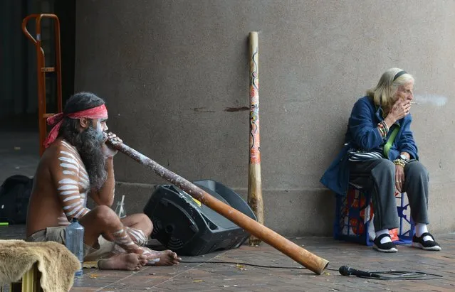 An elderly woman smokes a cigarette whilst an Aboriginal man plays a didgeridoo in Circular Quay in Sydney, Australia on April 4, 2017. (Photo by Peter Parks/AFP Photo)