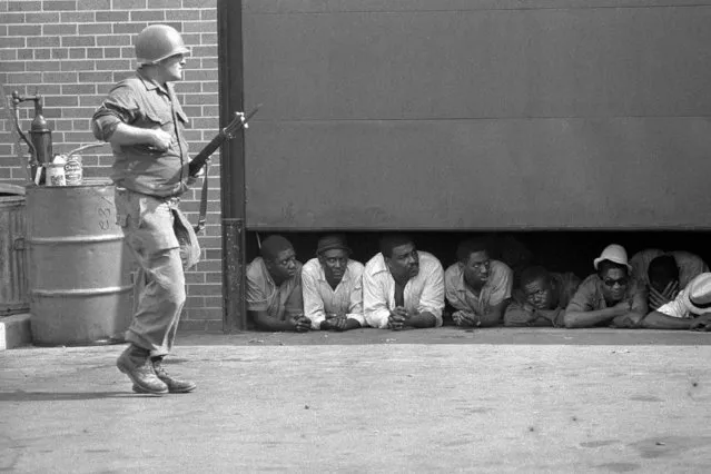 In this July 26, 1967 file photo, an Army soldier stands guard as men captured in the vicinity of the 10th Police Precinct in Detroit peer from under a garage door awaiting transfer. The precinct building came under fire in daylight hours and an Army force, using armed personnel carriers and tanks came to the police station. (Photo by AP Photo)