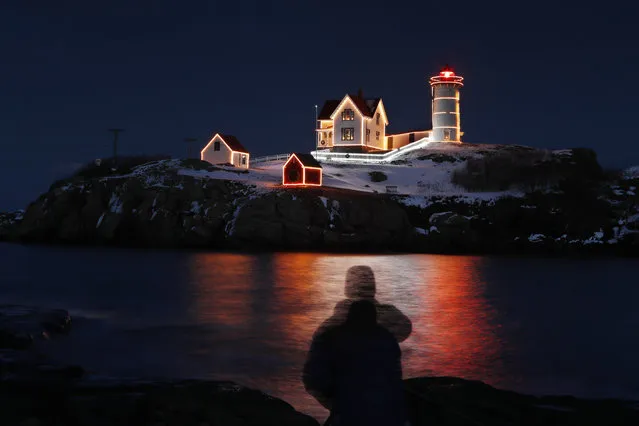 In this long exposure shot, holiday lights reflect in the waters off Nubble Light, silhouetting photographer Michael Blanchette of Bedford, N.H., Monday, December 11, 2017, in York, Maine. The beacon on top of the lighthouse has been helping mariners navigate the coastal waters of southern Maine since 1879. The seasonal decorations will stay lit through Jan. 1. (Photo by Robert F. Bukaty/AP Photo)