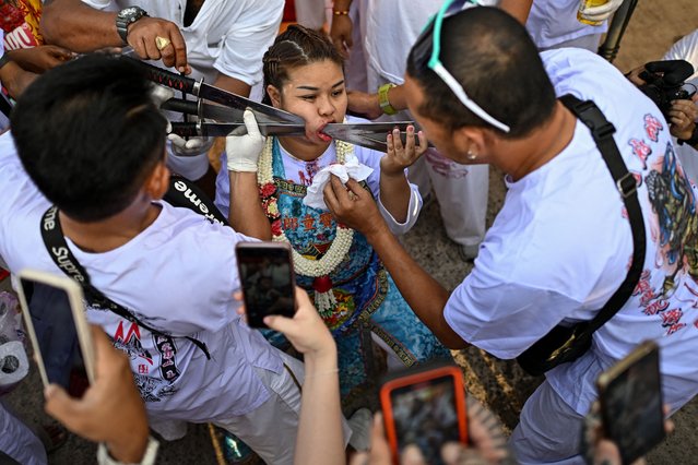 Sirinnicha Thampradit, a devotee of the Jor Soo Gong Naka shrine, has swords pierced through her cheek as she takes part in a procession during the annual vegetarian festival in Phuket on October 4, 2024. Soon after sunrise in the heart of the old town of the Thai island of Phuket, a swordsman chants an incantation and slowly slides the first of three blades into the right cheek of the woman who sits calmly below him. (Photo by Manan Vatsyayana/AFP Photo)