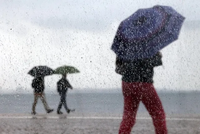 People, photographed through a window with raindrops, shelter under umbrellas against the rain as they walk near the Tagus riverbank in Lisbon, Wednesday, April 8, 2015. The city promenade is a popular riverside walking path where people stroll or enjoy fishing. (Photo by Francisco Seco/AP Photo)