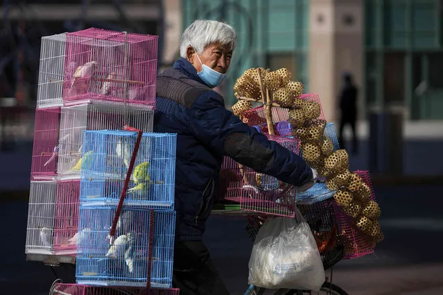 A vendor wearing a face mask to help protect from the coronavirus rides on a bicycle with cages of pet birds for sale along a street in Beijing, Tuesday, February 22, 2022. (Photo by Andy Wong/AP Photo)