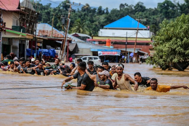 People holding a rope wade across flood water after the Kobe River overflowed following heavy rains in Lukulamo, North Maluku, on July 22, 2024. (Photo by Azzam Risqullah/AFP Photo)