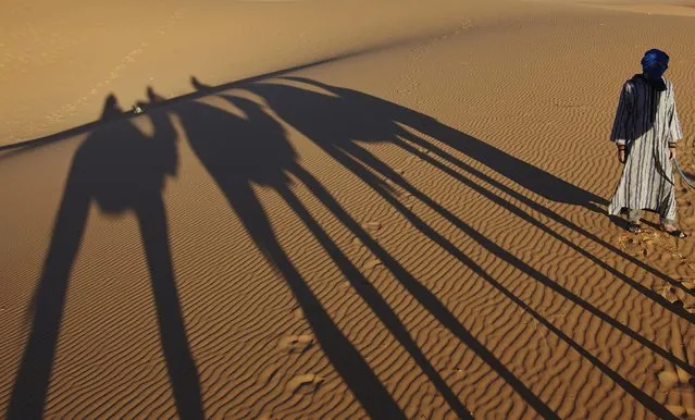 A camel vendor walks near the shadows of tourists riding camels, in Merzouga, Morocco along what is called the route of a thousand kasbahs in the Atlas Mountains, Sunday, March 5, 2017.  (Photo by Abdeljalil Bounhar/AP Photo)