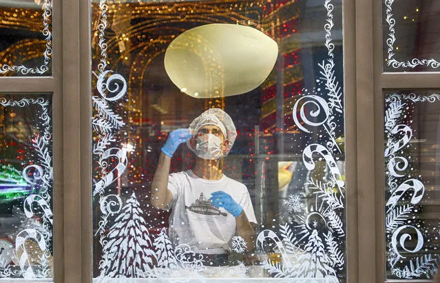 A cook in a face mask is seen making a pizza through a window glass decorated with Christmas ornaments in a city pizzeria in Kyiv, Ukraine, Monday, January 11, 2021. The country of 42 million is recording about 9,000 new COVID-19 infections a day and more than 19,500 people have died. Ukraine imposes a wide-ranging lockdown beginning Friday, closing schools and entertainment venues and restaurant table service through to Jan. 25. (Photo by Efrem Lukatsky/AP Photo)