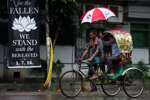 A man using a JICA emblazoned umbrella rides the back of a cycle rickshaw past a banner that reads “We stand with the bereaved” in a street near an upscale restaurant which was the site of a bloody siege that ended in the death of seventeen dead foreigners and five Bangladeshis, including two policemen, in Dhaka on July 5, 2016. Bangladesh's home minister said the attackers who stormed the upmarket cafe on the night of July 1, taking dozens of diners hostage and killing 20, were highly educated and from wealthy families. (Photo by Roberto Schmidt/AFP Photo)