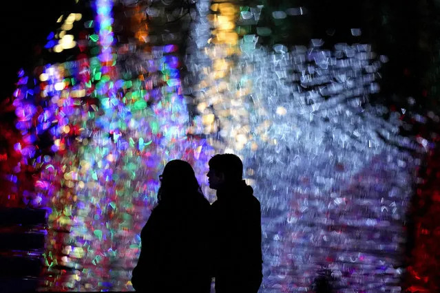People are silhouetted against Christmas lights reflected off a pond as they visit a park, Sunday, December 4, 2022, in Lenexa, Kan. (Photo by Charlie Riedel/AP Photo)