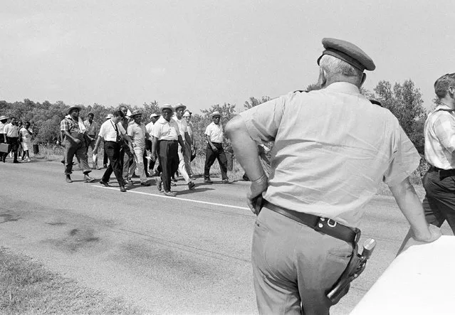 A Mississippi Highway Patrolman leans against a parked car along U.S. 51 as the front rank of marchers on the Memphis to Jackson trek approaches across the highway near Coldwater, Mississippi on June 9, 1966. Nobel Prize winner Dr. Martin Luther King (light trousers) and other civil rights leaders lead the march. (Photo by AP Photo)