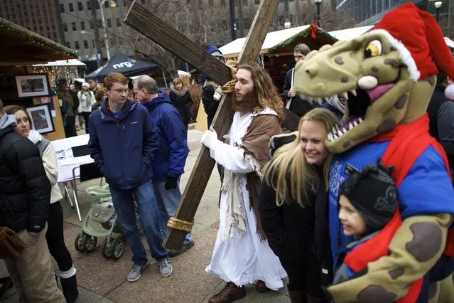 While visitors pose for photographs with a Santa Claus dinosaur,  Michael Grant, 28, “Philly Jesus”, walks through the Christmas Village in LOVE Park towards the nativity scene after carrying this 12 foot cross 8 miles through North Philadelphia to Center City as part of a Christmas walk to spread the true message of the holiday in Philadelphia, Pennsylvania December 20, 2014. (Photo by Mark Makela/Reuters)
