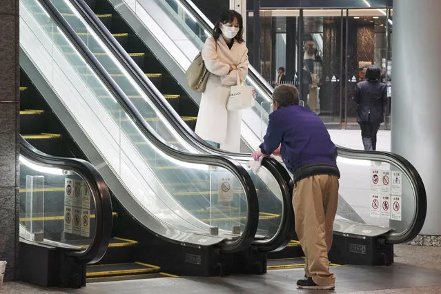 A woman wearing a mask as a precaution against a new coronavirus passes by a worker cleaning escalator handrails at a business building in Tokyo Monday, March 9, 2020. (Photo by Eugene Hoshiko/AP Photo)