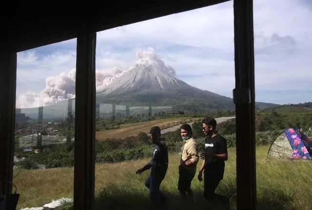 People walk past by as Mount Sinabung spews volcanic materials during an eruption in Karo, North Sumatra, Indonesia, Thursday, March 11, 2021. The 2,600-meter (8,530-feet) volcano unleashed an avalanche of searing gas clouds flowing down its slopes during eruption on Thursday. No casualties were reported. (Photo by Binsar Bakkara/AP Photo)