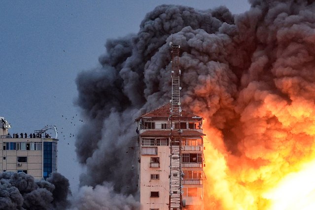 People standing on a rooftop watch as a ball of fire and smoke rises above a building in Gaza City on October 7, 2023 during an Israeli air strike that hit the Palestine Tower building. One month after Israel was wracked by Hamas attacks, life has been upended for both the Palestinians and Israel after it launched a war of reprisal in the Gaza Strip. The October 7 attacks by Hamas militants who stormed across from Gaza and struck kibbutzim and southern Israeli areas killed 1,400 people, mostly civilians, and deeply scarred the nation. The health ministry in Hamas-run Gaza says nearly 9,500 have been killed, two-thirds of them women and children, and mostly civilians. (Photo by Mahmud Hams/AFP Photo)