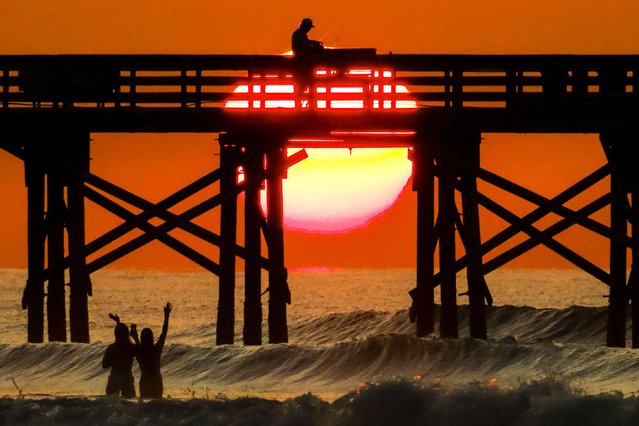 A man fishes from the Isle of Palms pier as young girls record a video silhouetted by the sunrise over the Atlantic Ocean along Front Beach, August 18, 2025 in Isle of Palms, South Carolina. High waves and potential storm serge is expected later in the week as Hurricane Erin, a category 4 storm passes off the coast. High waves and potential storm serge is expected later in the week as Hurricane Erin, a category 4 storm passes off the coast. (Photo by Richard Ellis/Alamy Live News)