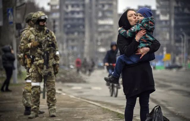 A woman holds and kisses a child next to Russian soldiers in a street of Mariupol on April 12, 2022, as Russian troops intensify a campaign to take the strategic port city, part of an anticipated massive onslaught across eastern Ukraine, while Russia's President makes a defiant case for the war on Russia's neighbour. (Photo by Alexander Nemenov/AFP Photo)