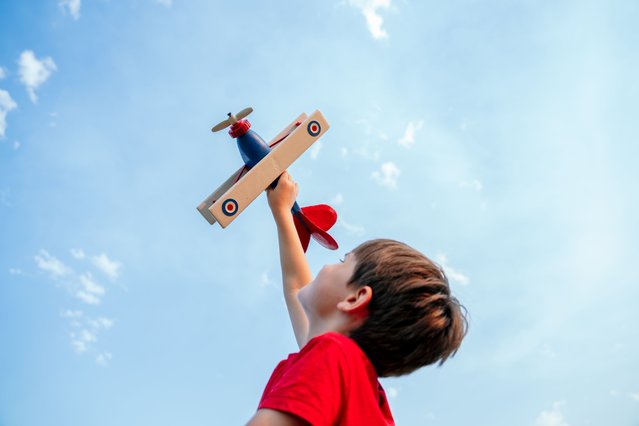 Low angle view of little boy flying in the blue sky a handmade wooden airplane. Childhood and travel concept. (Photo by Vera Vita/Getty Images)