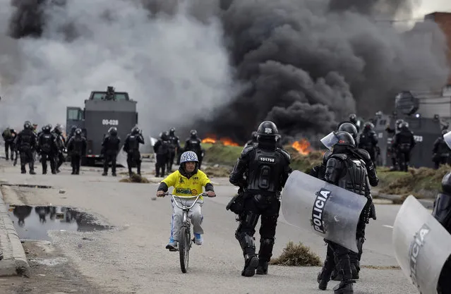 A man rides his bicycle past police clashing with striking truckers in Bogota, Colombia, Wednesday, July 20, 2016. Hundreds of truckers clashed with police on Wednesday on the 41st day of their strike which has made food scarce in some areas of the country. Truckers are demanding a higher price for freight, lower fuel prices and fewer license regulations for cargo. (Photo by Fernando Vergara/AP Photo)