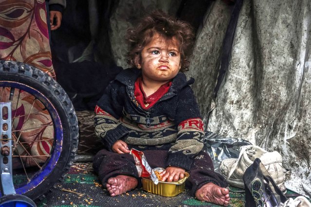 A displaced Palestinian child eats food from a box while sitting in a makeshift tent at a camp beside a street in Rafah on March 13, 2024, amid ongoing battles between Israel and the Hamas militant group. The Israel-Hamas conflict raging since October 7 has caused mass civilian deaths, reduced vast areas to a rubble-strewn wasteland and sparked warnings of looming famine in the Palestinian territory of 2.4 million people. (Photo by Mohammed Abed/AFP Photo)