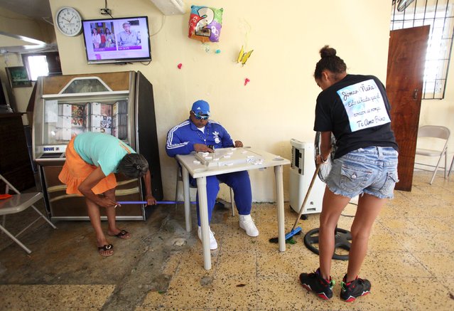 The mother (L) of Jomar Aguayo and a friend sweep the floor around the table where the dead body of Jomar is seated with domino tiles in San Juan, October 19, 2015. Aguayo's family decided, with the help of a funeral home that specializes in embalming techniques, to have him sit at a table at his mother's bar for the wake. Aguayo was 23 years old when he was shot dead during a shootout, according to local media. (Photo by Alvin Baez/Reuters)
