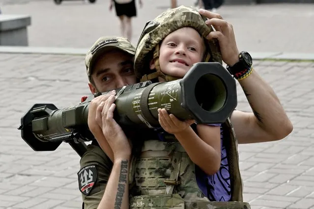 A Ukrainian serviceman helps a boy wearing body armour to hold an AT4 Swedish man-portable anti-tank weapon at a volunteers point in the centre of Kyiv on August 10, 2023, amid Russian invasion in Ukraine. (Photo by Sergei Supinsky/AFP Photo)