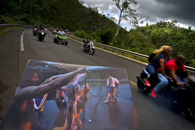 A printed photograph taken on September 28, 2017 shows people bathing on the highway after Hurricane Maria destroyed people's homes, held up at the same spot of the highway where motorcyclists ride past in Naranjito, Puerto Rico, May 27, 2018. Thanks to the owners of the land alongside the highway, creek water was piped to the side of the road for people without water to use for bathing, washing clothes and dishes. (Photo by Ramon Espinosa/AP Photo)