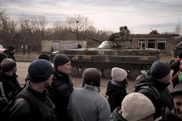 Residents lining up for aid watch as Ukrainian soldiers ride atop a tank in the town of Trostsyanets, Ukraine, Monday, March 28, 2022. Trostsyanets was recently retaken by Ukrainian forces after being held by Russians since the early days of the war. (Photo by Felipe Dana/AP Photo)