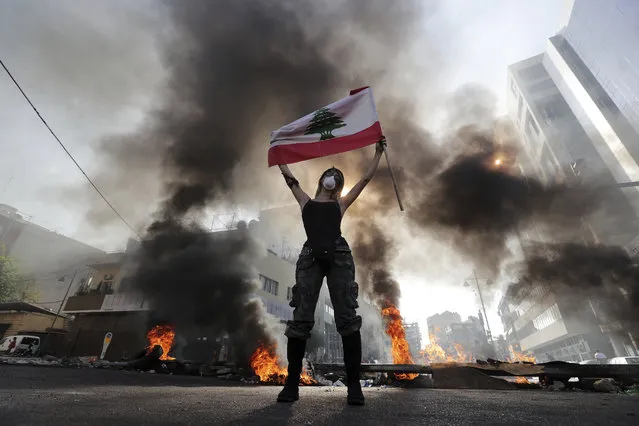 An anti-government protester holds a Lebanese flag while standing in front burning tires that block a road in the town of Jal el-Dib, north of Beirut, Lebanon, Wednesday, November 13, 2019. Lebanese protesters blocked major highways with burning tires and roadblocks on Wednesday, saying they will remain in the streets despite the president's appeal for them to go home. (Photo by Bilal Hussein/AP Photo)