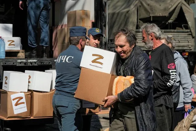A man carries bread and boxes with the letter Z, which has become a symbol of the Russian military, and a hashtag reading “We don't abandon our own”, as other local civilians gather to get humanitarian aid, bread and water distributed by Donetsk People Republic Emergency Situations Ministry in Mariupol, in territory under the government of the Donetsk People's Republic, eastern Ukraine, Friday, April 29, 2022. This photo was taken during a trip organized by the Russian Ministry of Defense. (Photo by AP Photo/Stringer)